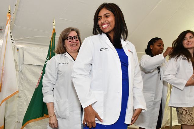 Photo: A woman with a blue dress and short, dark hair wears a white lab coat in front of a woman with short, light brown hair at a white coat ceremony. To the right of the two individuals in the center are two other individuals in the exact position, both smiling as the one behind helps the one in front don her coat. They are standing on a covered stage.