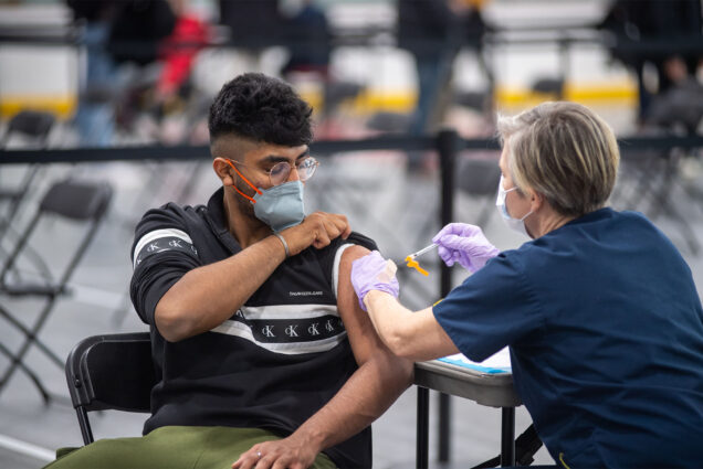 Photo: A person in a mask and glasses raises the sleeve of their shirt to receive a shot from a healthcare professional