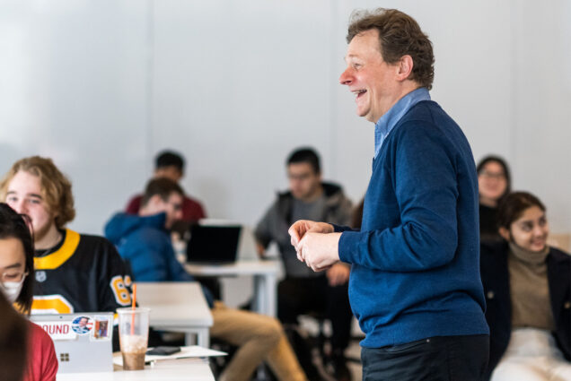 Photo: A man in a blue sweater with red hair laughs as he teaches a classroom full of college students