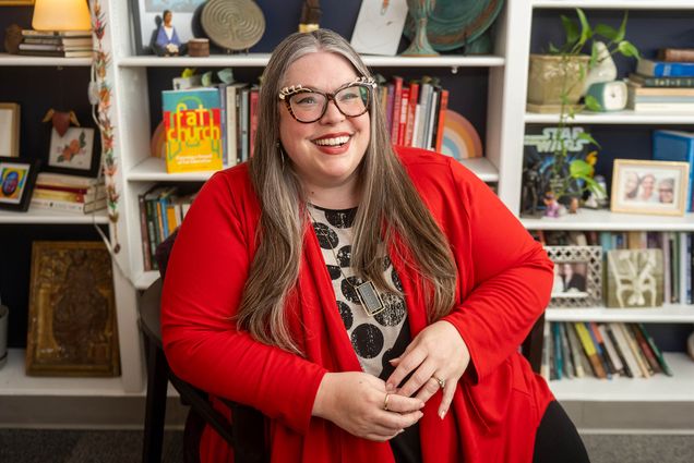 Photo: A person in an orange cardigan with glasses poses for a picture in front of a wall of books