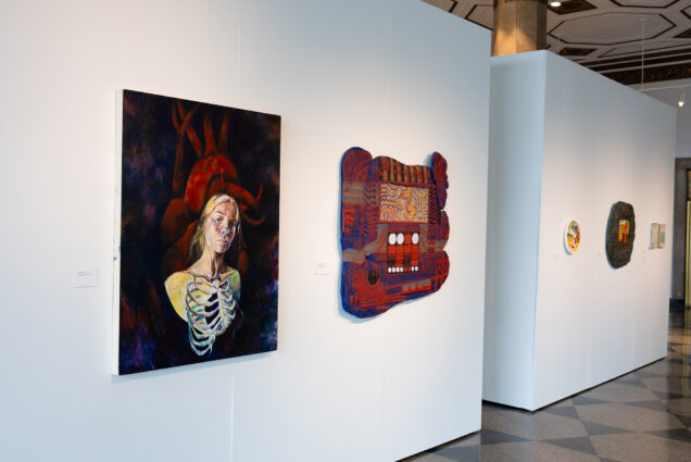 Photo: large canvases featuring various paintings are shown on display on large, white gallery walls.Art closest to left shows a portrait with a skeleton overlay and next to it on the right is an abstract work of red and blue.