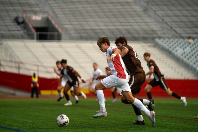 Photo: College soccer players race each other towards a soccer ball on a large green field in front of a row of metal bleachers