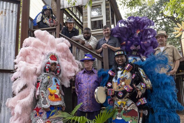 Photo: A group of people wearing bright, flamboyant Mardi Gras Indian outfits stand and pose in front of a large stoop. Large pink and purple feathers are showcased on outfits and headdresses.