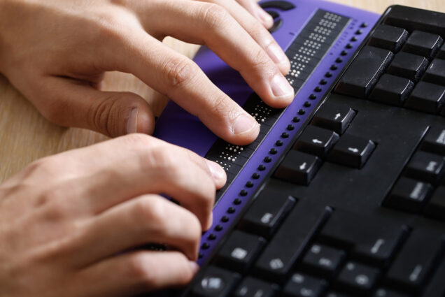 Photo: A set of hands use a computer with a braille computer display and a computer keyboard.
