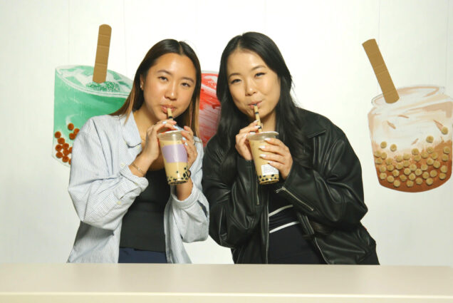 Photo: Sharlene Chang (SHA’24) and Irene Kim (COM’23), two young Asian women sipping boba teas, smile and pose resting together. They sit in front of a backdrop of large boba tea recreations made out of cardboard and puff balls.