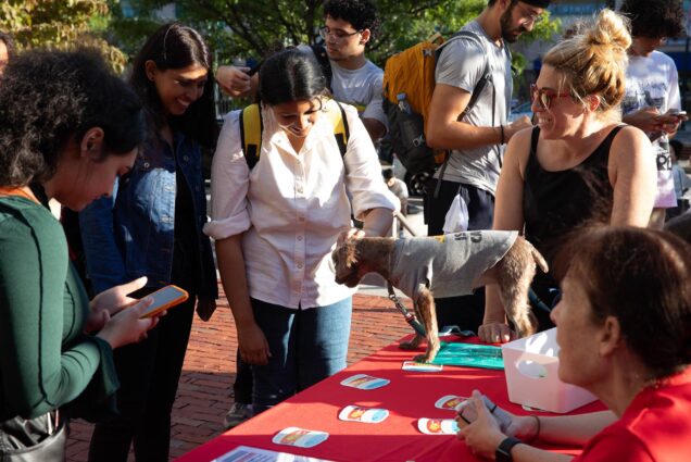 Photo: Various students smile as they pet a small dog wearing a grey shirt that stands atop a red table. A woman to the dog's right smiles and looks on as various stickers and pamphlets are on display on the table.