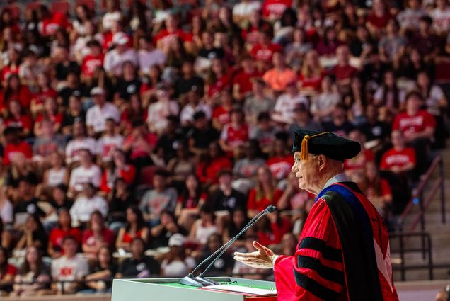 Photo: Interim president Ken Freeman speaks at a podium to a large crowd of students at the 2023 Freshman Matriculation ceremony. He wears a red and black ceremonial rove and black cap as students watch on.