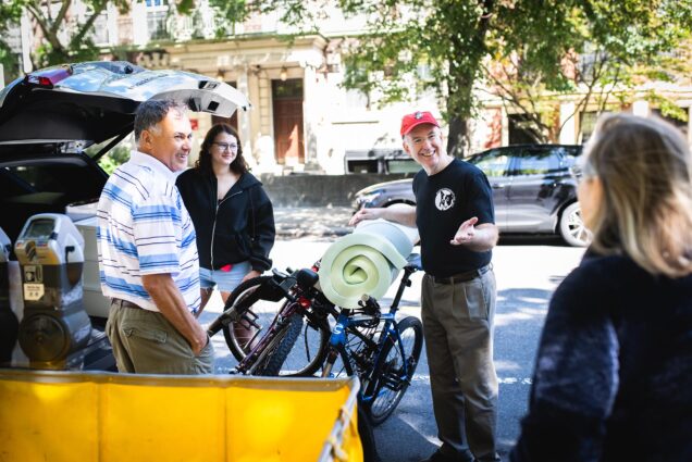 Photo: Kenneth Freeman, an older white man wearing a red BU cap, black shirt, and khaki pants, smiles as he helps unload a bike from the back of a car. A student and her parents help and smile as they chat with him on the sunny street.