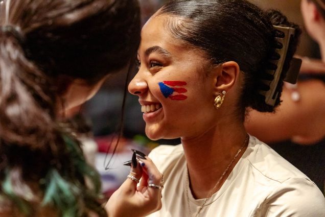 Photo: A young Latina woman looks to the left and smiles as another person paints a Puerto Rican flag on her cheek.