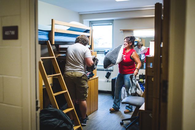 Photo: A woman wearing a red shirt and jeans laughs as she holds a large pillow in her hands. The person in front of her hold various clothes and places them away in a dresser in front of them/