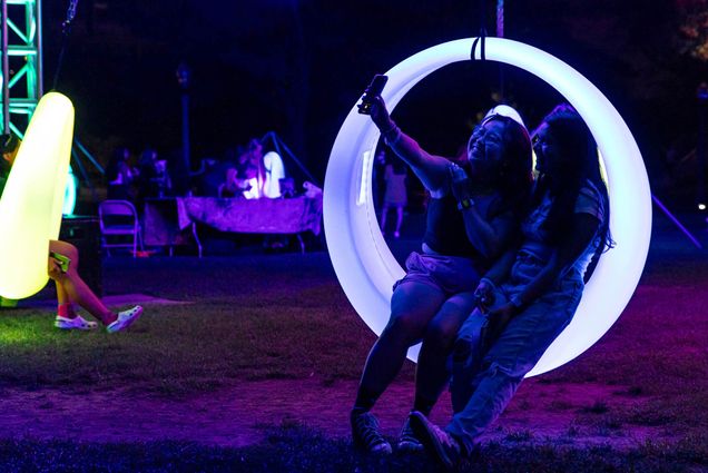 Photo: Two students sit on a large swing made up of a ring of light and pose together as they take a selfie. The scene is lit up by the swing and similar swings around them during the night.