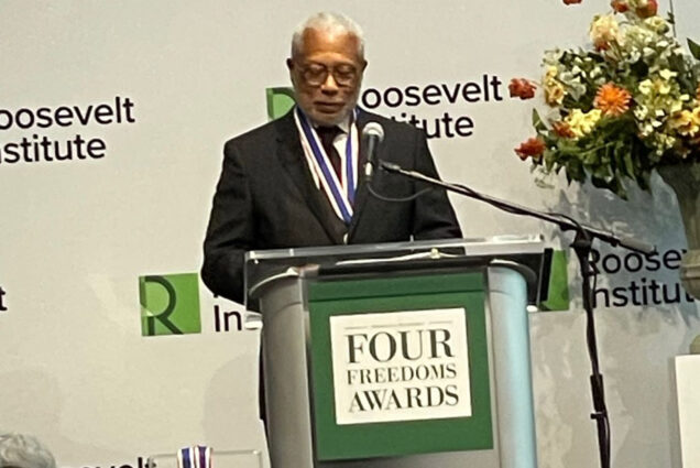 Photo: Dr. Walter Earl Fluker, an older Black man with white-grey hair and wearing a black suit and large medala round his neck, looks down as he stands and speaks at a podium. Sign on podium and backdrop behind him both read "Four Freedoms Awards".