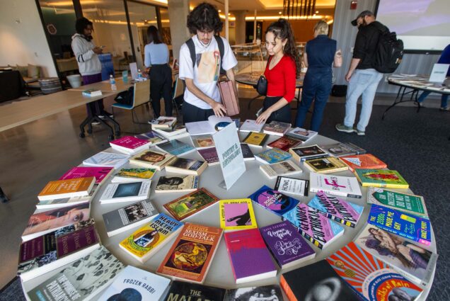 Photo: Two students look through a neatly arranged display of Poetry and Anthology books laid out on a round white table. Other students and people can be seen looking around other tables with similarly arranged displays of books.