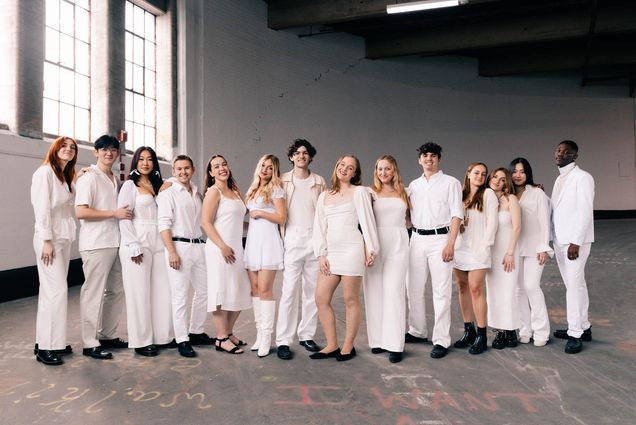 Photo: Members of BU's acapella group, Trebelmakers, stand in a land for a group shot. They are in a warehouse and all members are wearing personal variations of a white outfit. They are stand, candidly.