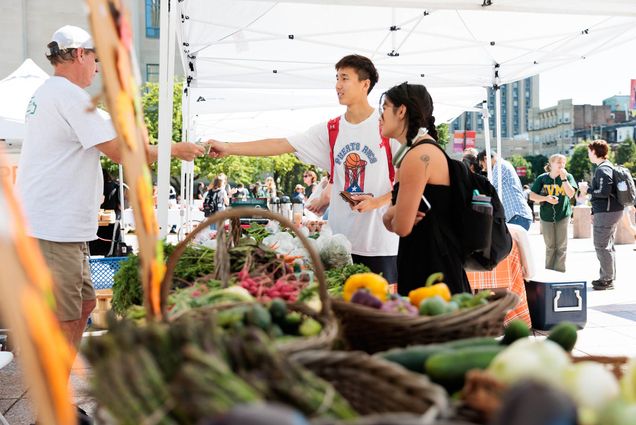 Photo: Two students with dark hair pay a man at a farmers market stand. They are under a white tent, the day is bright, and various vegetables lay in brown wicker baskets.