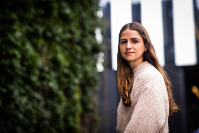 Photo: A woman with long, light brown hair and a cream sweater looks over her shoulder for a portrait shot. On her left, there is a contrasting greenery and behind, blurred out beginnings of a building.