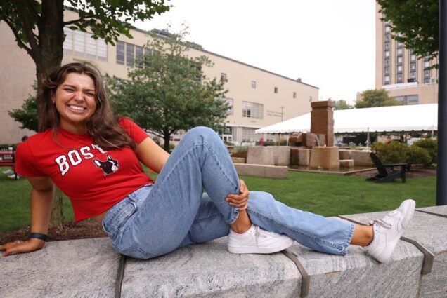 Photo: A college student wearing jeans and a red t shirt leans back on a stone wall on BU's campus.