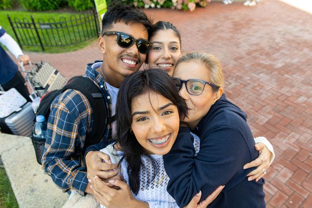 Photo: A group of individuals hugging and embracing each other as the look up at the camera. A woman with braces smiles, behind her a man with a flannel and glasses holds her shoulder and smiles, next to him, another woman with her hair pulled back smiles with their heads next to each other. On the right, a woman with glasses nuzzles into the embrace, peaking a look at the camera with her one visable eye.