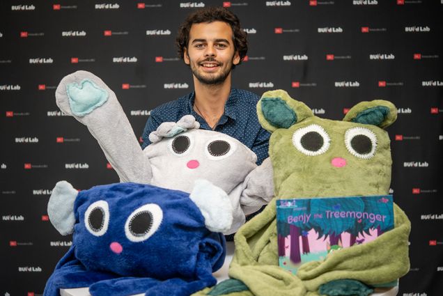 Photo: A man with a button-up shirt sits behind three stuffed creatures, all with huge eyes and long limbs. They are characters from the children book that one of the creatures holds, a book for sustainability for kids.