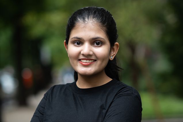 Photo: A young woman with her arms crossed. smiling for the camera. She has her hair pulled back into a ponytail and she wears a black t-shirt and dark jeans. The blurred background behind her is of sidewalk and green foliage.