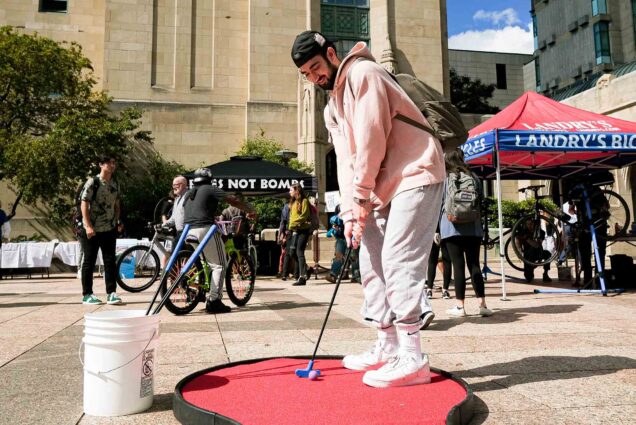 Photo: A white man in a pink sweatshirt and gray sweatpants stands on mock pad of a golfing turf. The day is bright and there are different tents of activities around him.