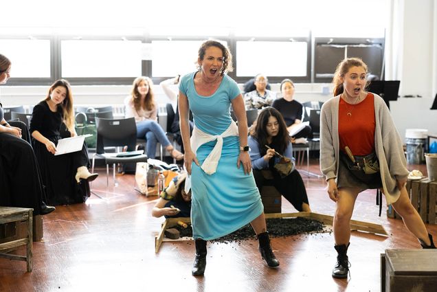 Photo: A woman in a blue dress next to a woman in a red shirt who appears to be yelling performs in front of a room of students watching from chairs in the background