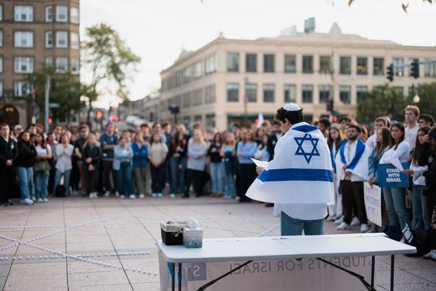 Photo: Students gather in front of Marsh Plaza on October 11th for a vigil organized by BU Students for Israel in partnership with BU Hillel. A young man wearing the white and blue Israeli flag around his shoulders and white yarmulke, stands at the front and center of the crowd as he reads a speech. He stands in front of a white plastic table as a large crowd holding various signs of support for Israel give him space and look on.