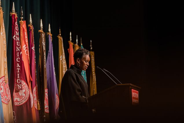 Photo: Dr. Melissa L.Gilliam, a Black woman with a short afro and wearing a black turtleneck sweater, speaks into a microphone on a wooden podium. Sign on podium reads "Boston University". Behind her, a row of colorful flags are placed.