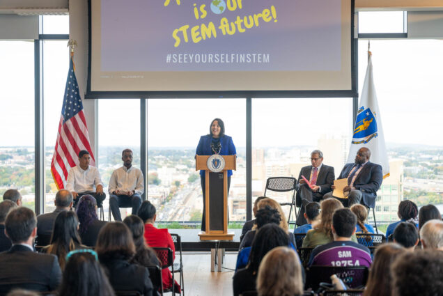 Photo: Lt. Governor Kim Driscoll speaks at the kickoff of the MA STEM Week. A tan woman wearing a blye blazer and black pants smiles and speaks at a wooden podium set up on a small stage. The stage is placed in front of large windows letting in lots of light. On her left are two students wearing long-sleeved, white collared shirts and slacks and to her left sits , a white man wearing glasses and a black suit, Massachusetts Secretary of the Executive Office of Education Patrick Tutwiler, a Black man wearing glasses and a black suit.