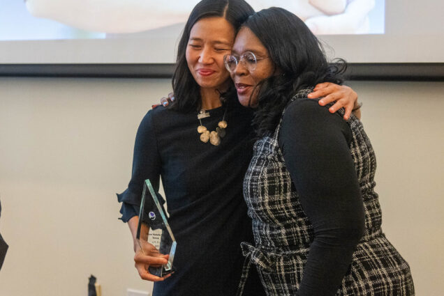 Photo: (left) Boston Mayor Michelle Wu embraces Monalisa Smith (right). Wu, an Asian woman with black hair and wearing a black dress, smiles and rests her right arm over the shoulders of Smith. In her left hand she holds a small, glass trophy. Smith, a Black woman wearing glasses, a long-sleeved black shirt, and grey plaid dress, leans into Wu's embrace and smiles. they both pose for another camera.