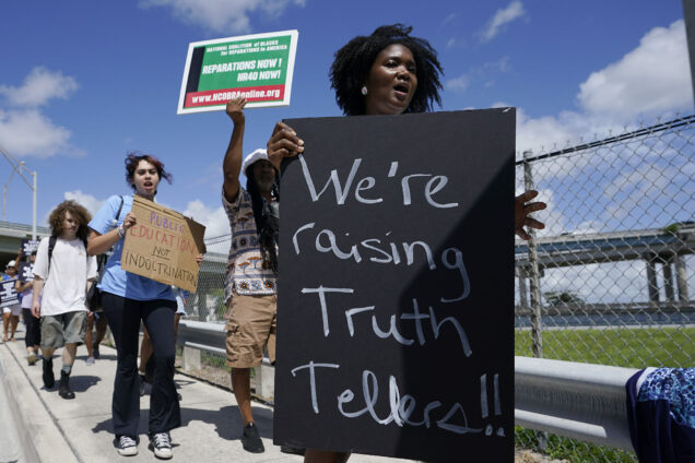 Photo: People carry signs during the "Teach No Lies" march to the School Board of Miami-Dade County to protest Florida's new standards for teaching Black history. A Black woman in the front holds a large black sign with the words "We're raising truth tellers!" in white chalk.