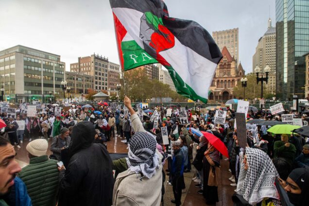 Photo: Stop the Gaza Genocide Protest in support of Palestine in Copley Square October 16. A crowd of people march and chant slogans on a gloomy day in Boston's Copley Square. One person holds and waves a large Palestinian flag in the foreground.