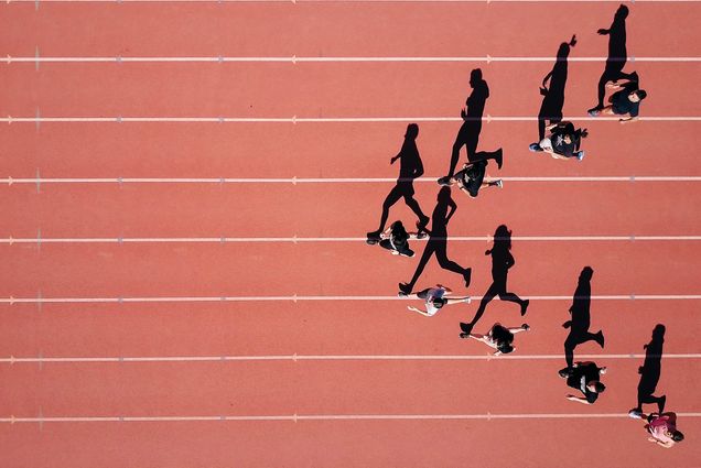 Photo: A group of runners running in a arrow formation on a outdoor track. The photo is from a birds eye view.