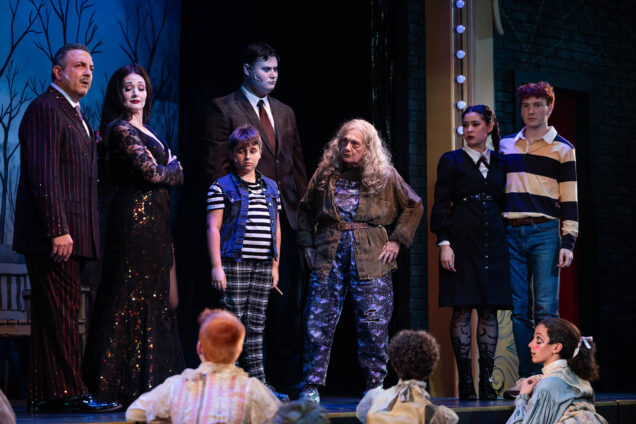 Photo: From (left to right) Luis Negron playing Gomez Addams, Aimee Doherty as Morticia Addam, Jack Baumrind as Pugsley Addams, Sam David Cohen as Lurch, Jane Stabb as Grandma, Emilia Tagliani and Timothy Bevens as Lucas Beineke, act out a scene during a dress rehearsal for the play, The Addams Family at Wheelock Family Theatre. A group of people dressed in spooky black, emo attire act out a scene on stage as the rest of the cast looks on.
