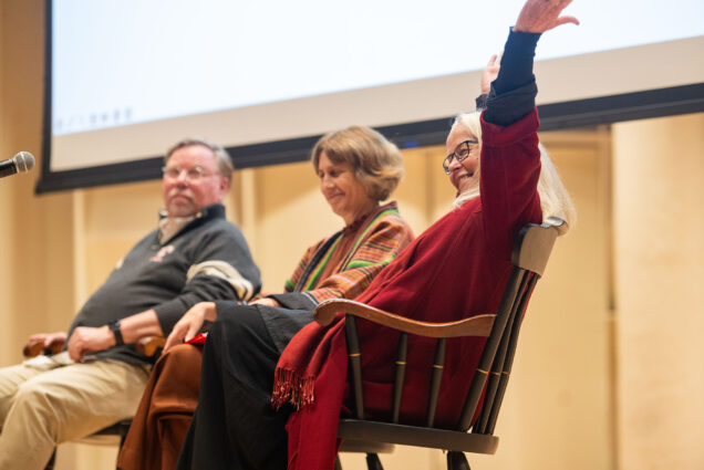 Photo: (from left) Gregory Melchor-Barz, Dana Clancy, and Susan Mickey sit in chairs on a stage and look towards an unseen audience. Mickey waves a hand high in the air and smiles towards the audience.