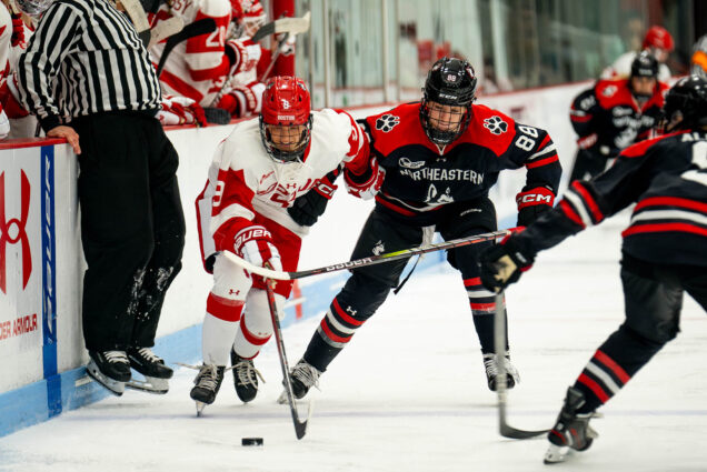 Photo: A woman in a red and white BU ice hockey uniform fights for the puck against two opposing Northeastern players dressed in black and red uniforms. A referee can be seen to the left of them leaning out of the way of play against the ice rink barrier.