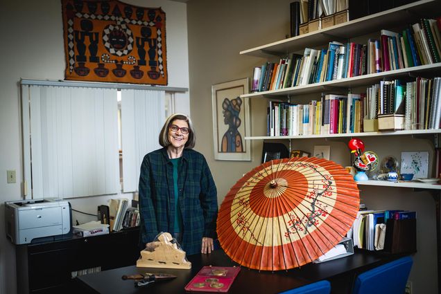 Photo: A photo of Marianne Farkas in her office, smiling for the photo. She wears a plaid button up with a blue shirt underneath. Her hair is white stripes with the rest, black, with rectangle glasses. The objects highlighted in her office are around her.