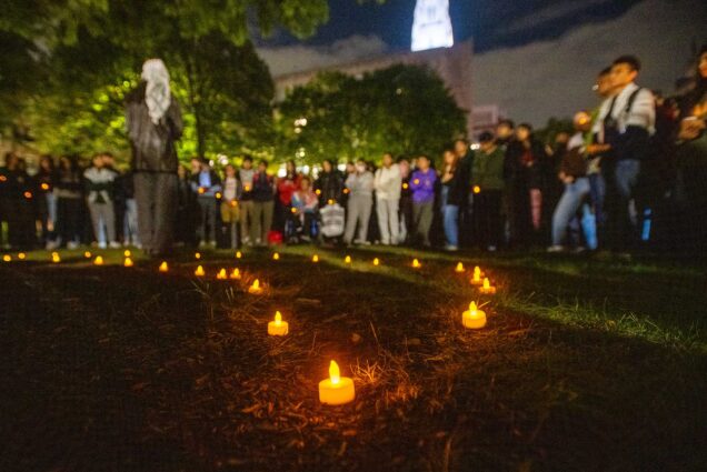 Photo: A group of people and students stands as a perosn in the center gives a speech. The person in the center faces away from the camera and faces of crowd are blurred for privacy. Electric candles are shown placed on the ground during the clear evening.