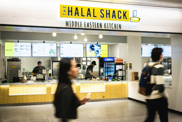 Photo: Exterior shot of the Halal Shack in BU's student union. A food court restaurant with a bright interior and large yellow sign that reads "The halal shack" in black letters is shown. Blurred students are shown walking by in the foreground as workers make bowls behind the counter.