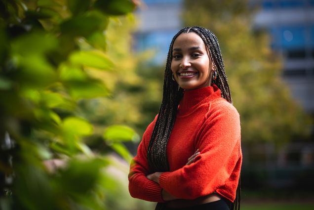 Photo: Hermela Haile, a young Black woman with long brown and light brown box braids and wearing a red orange turtleneck and black pants, smiles and poses with arms crossed in front of lush greenery.