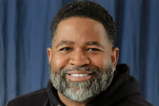 Photo: Jamil Smith, a man with a beard, smiles for the camera. He is wearing a black hoodie and is in front of a blue curtain background.