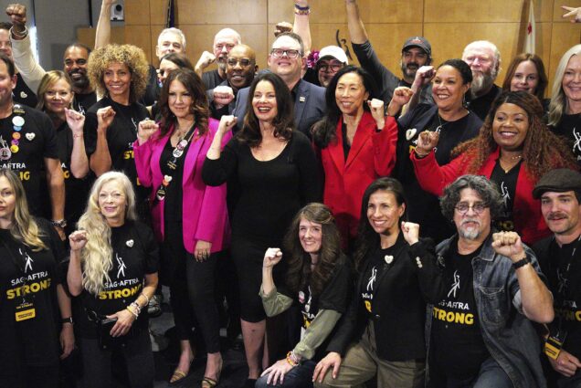 Photo: GFran Drescher, center, joined with National Executive Director and Chief Negotiator Duncan Crabtree-Ireland, and the TV/Theatrical Negotiating Committee members posing with one fist up in the air. They stand in front of a wooden wall.