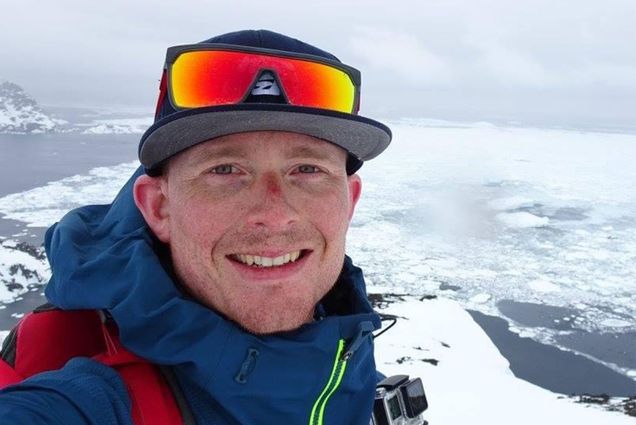 Photo: A White man wearing a hat and sunglasses stands atop a snowy mountain