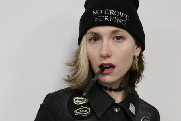 Photo: A woman with short blonde hair wearing a black beanie that says "No Crowd Surfing" and a jacket covered in pins
