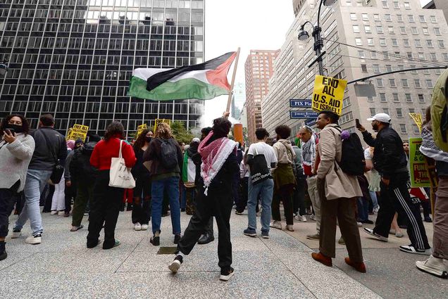 Photo: A group of protestors are shown standing in front of the Consulate General of Israel along Second Avenue, New York, NY. A person in the center holds a large Palestinian flag above the crowd.