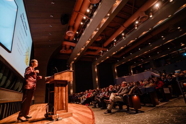 Photo: A woman in a suit stands at a podium in front of a crowded auditorium, giving a lecture