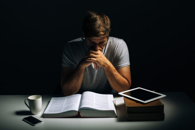 Photo: A stock image of a man studying with books, papers, and additional educational accessories spread out on a table. he is shadowed by a spotlight to his right, as he contemplatively looks over everything on the spread.
