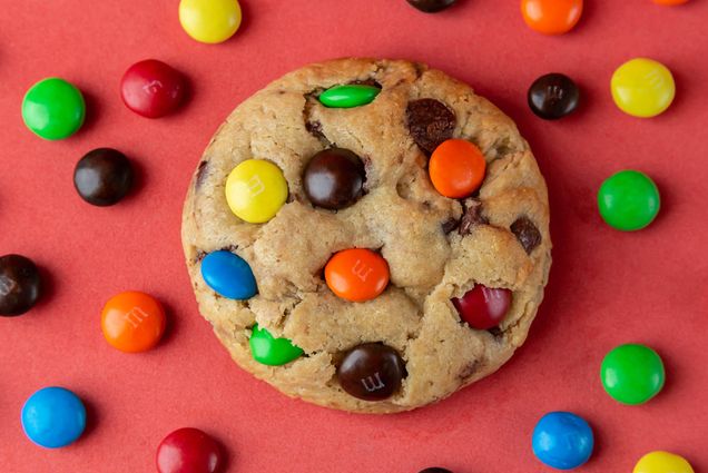 Photo: A stock image of a cookie with colorful candy-coated chocolates smushed on top. The cookie rests on a flat, light red background with various candy-chocolates scattered artistically around.