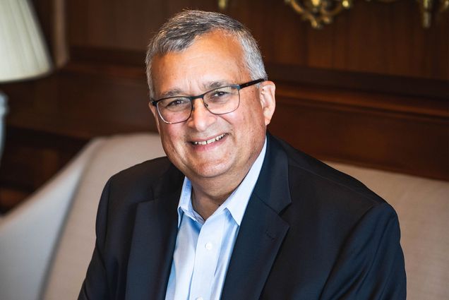 Photo: A middle aged man wearing glasses with a smile on his face sits in a high class couch, wearing a black suit and collared shirt.