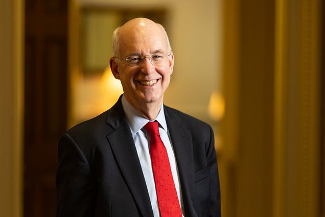 Photo: Kenneth Freeman, a white man wearing glasses, a light blue collared shirt, red tie, and black suit smiles and poses at 1 Silber Way in an elaborate hallway.